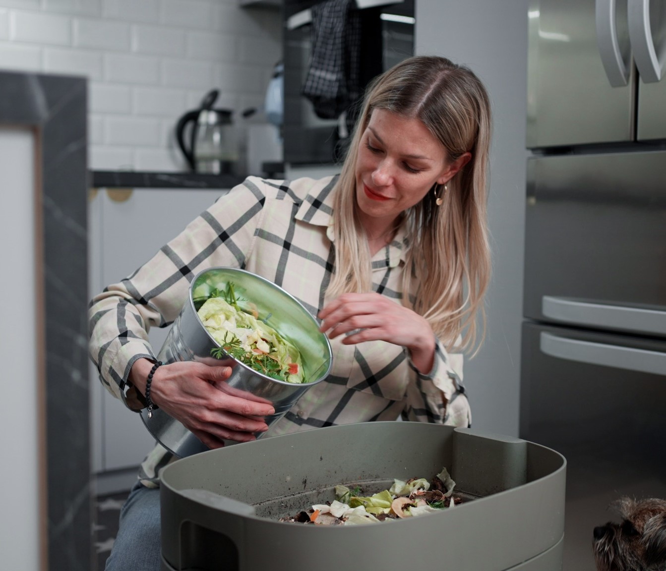 A woman in a modern kitchen empties a bowl of vegetable scraps into a countertop compost bin, while a small dog looks on nearby.